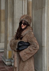 Manteau en fausse fourrure couleur fauve avec manches larges, complété par un chapeau assorti. Gants en cuir noir et pochette assortie. Des lunettes de soleil noires viennent parfaire le look.