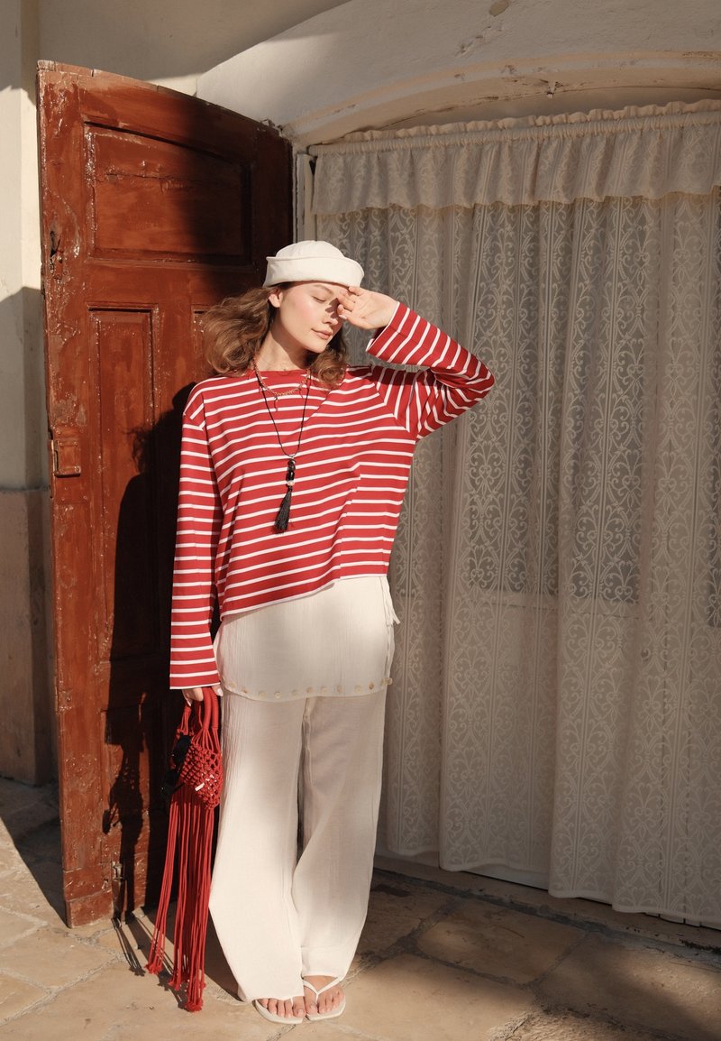 Femme en pantalon blanc et chemise rouge rayée portant un béret blanc, debout en se protégeant les yeux près d'une porte marron et de rideaux en dentelle.