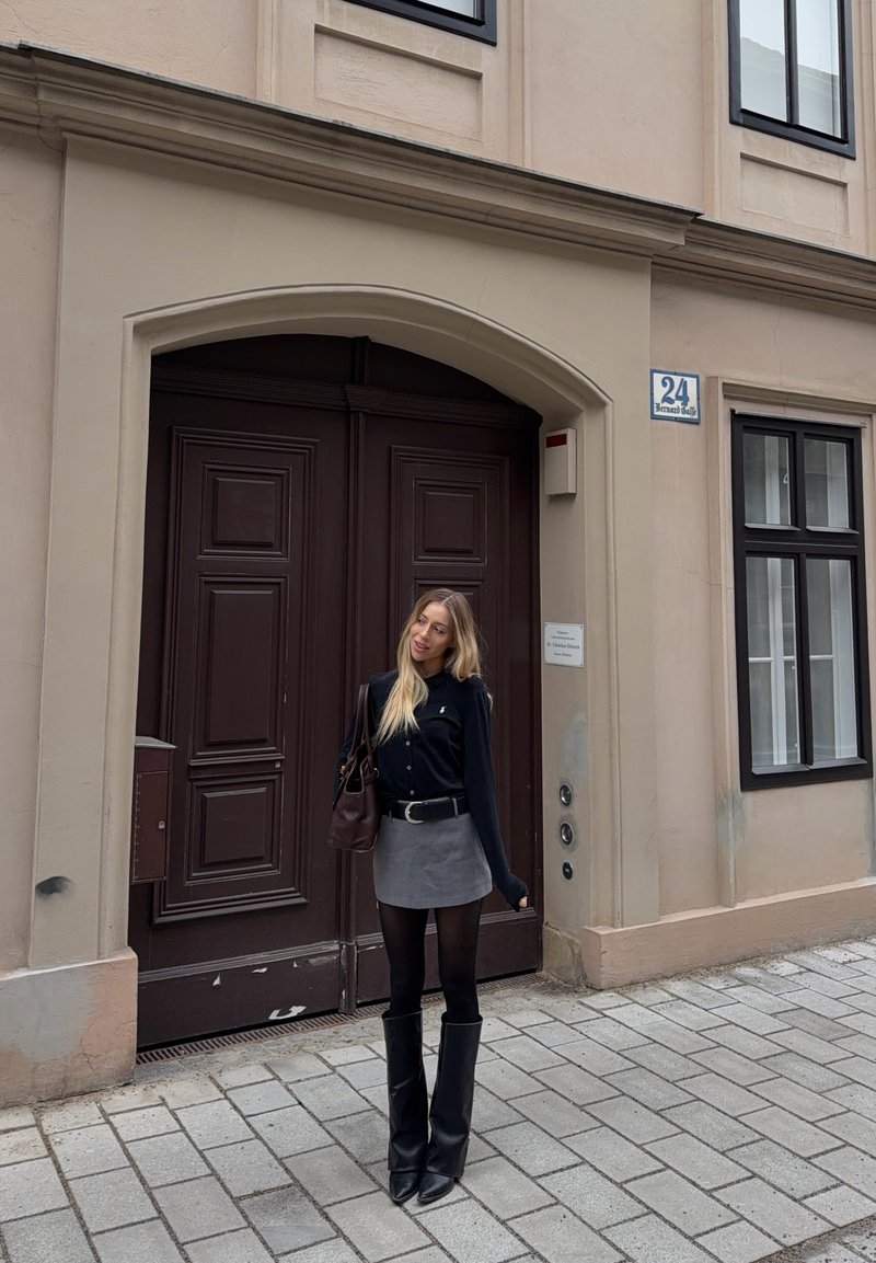 Jeune femme en pull noir, jupe grise, bottes noires hautes, debout sur une rue pavée devant une grande porte marron et un bâtiment beige.