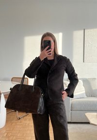 Schwarze Wildleder-Blazer mit Reverskragen und Knöpfen, kombiniert mit passenden weit geschnittenen Hosen. An der Seite wird eine schwarze Lederhandtasche gehalten.