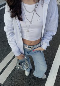 Γκρι φούτερ με φερμουάρ, λευκό crop top, ανοιχτού μπλε τζιν με φαρδιά πόδια και διακοσμητική ζώνη, στρωματοποιημένα ασημένια κολιέ, και τσαντάκι σε λευκή και γκρι παγιέτα.