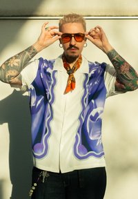 Košile s krátkým rukávem na knoflíky v bílé barvě s fialovým abstraktním vzorem, nošená s oranžovým šátkem, slunečními brýlemi a tmavými džínami.