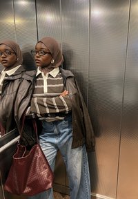 Brun læderjakke over en stribet brun og hvid skjorte, højtaljede denimjeans og en bordeaux vævet taske i en elevator.