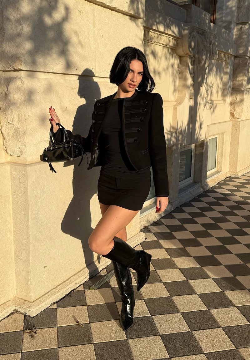 Femme en mini-robe noire, veste noire et bottes montantes appuyée contre un mur en pierre éclairé par le soleil sur un pavement à damier.