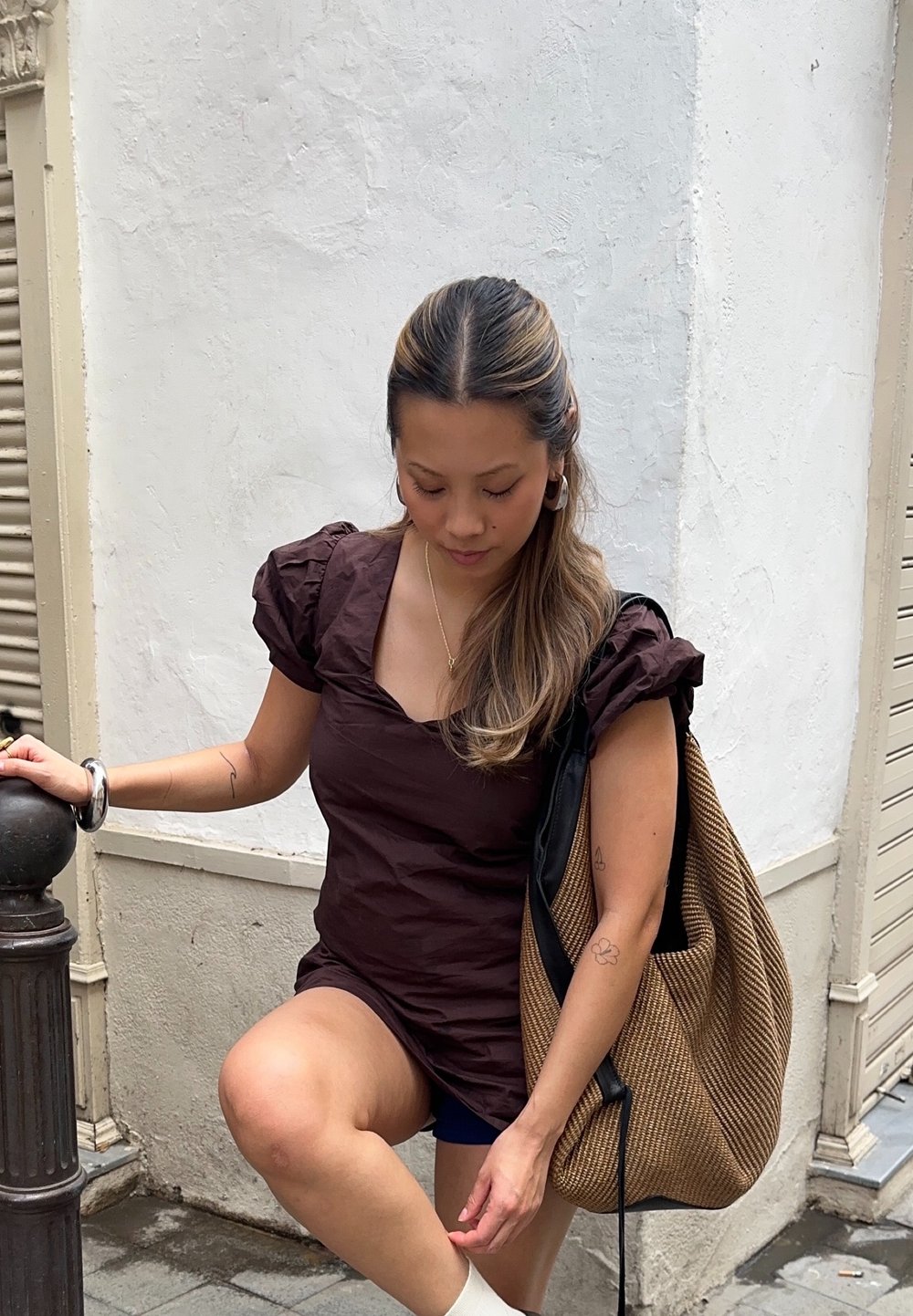 Haut marron à manches bouffantes avec un décolleté carré, associé à un short bleu marine. Tenant un grand sac brun texturé. Cadre de rue tranquille.