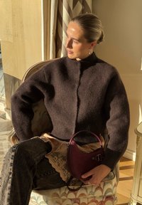 Femme aux cheveux tirés en arrière assise au soleil sur un fauteuil à motifs, portant un pull doux marron foncé, un jean, tenant un petit sac à main bordeaux.