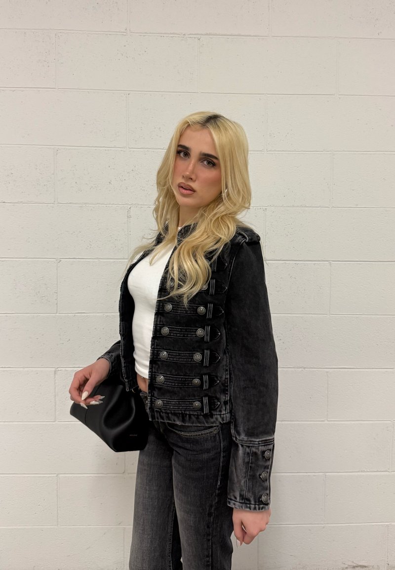 Jeune femme aux longs cheveux blonds portant une veste noire boutonnée, un haut blanc et un jean foncé, tenant un petit sac noir contre un mur blanc.