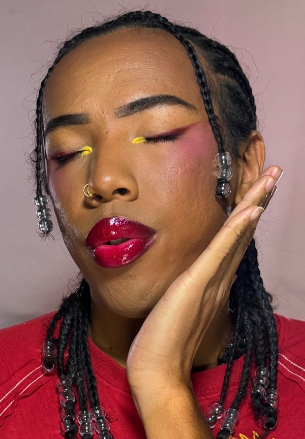 Personne aux cheveux tressés avec des perles claires, portant du rouge à lèvres rouge et un fard à paupières jaune, posant les yeux fermés et la main effleurant doucement le visage.