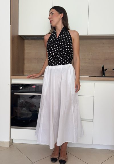 Mujer con top de lunares blanco y negro de tirantes halter y falda larga blanca, de pie en una cocina moderna, mirando hacia un lado.