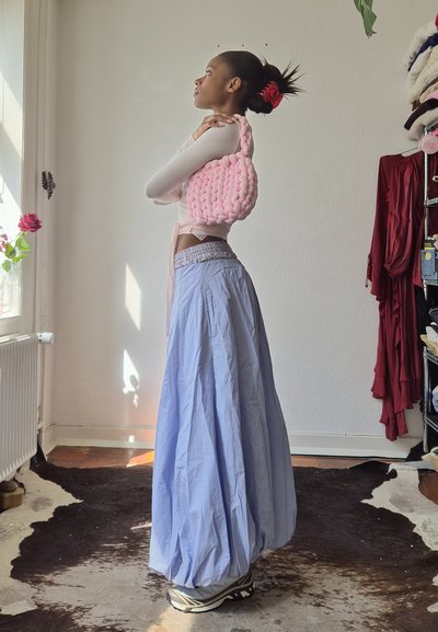 Mujer con el pelo corto recogido con una coleta roja sostiene un bolso rosa voluminoso sobre el hombro, lleva una falda larga azul y zapatillas deportivas.