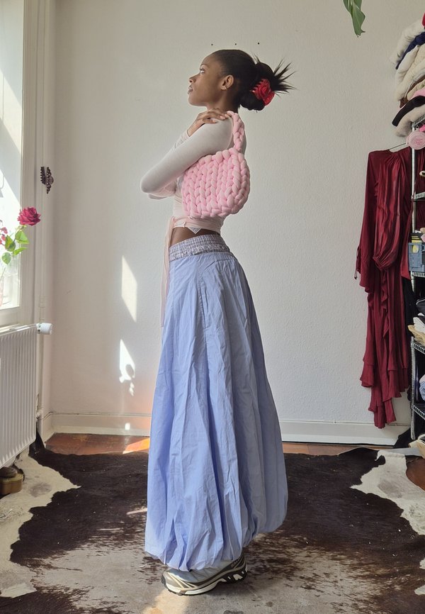 Kvinne med kort hår bundet med en rød scrunchie holder en stor rosa håndveske over skulderen, iført et langt blått skjørt og sneakers.