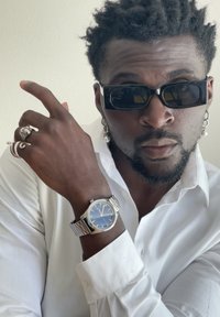 Sølv wristwatch med blå urskive, sølvrem og røde accenter. Hvid skjorte, sorte solbriller, flere ringe og perleøreringe.