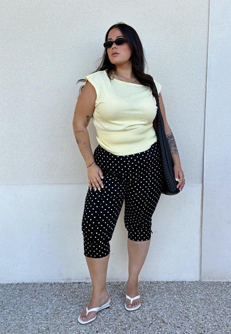 Femme aux longs cheveux foncés portant des lunettes de soleil noires, un haut jaune sans manches, un pantalon capri noir à pois, des sandales blanches et portant un sac noir.