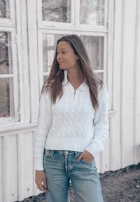 Pullover bianco a maglia con zip sul colletto, abbinato a jeans azzurri a vita alta, sullo sfondo di finestre in legno.
