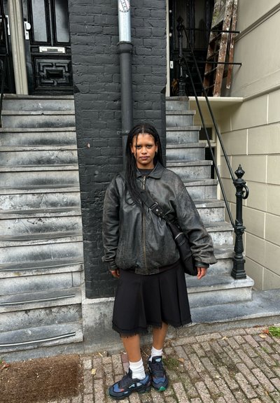 Chaqueta de cuero negra, falda negra y zapatillas con estampado. De pie contra una pared negra texturizada y escalones de piedra, llevando un bolso cruzado.