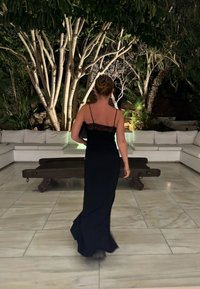 Vestido negro sin mangas con detalles de encaje en la parte posterior; tejido suave; hasta el suelo; combinado con un bolso negro en un entorno exterior bien iluminado.