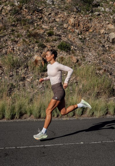 Mujer atlética corriendo por una carretera pavimentada junto a un terreno rocoso con hierba seca, vistiendo una camiseta ligera de manga larga, pantalones cortos oscuros y zapatillas de correr.
