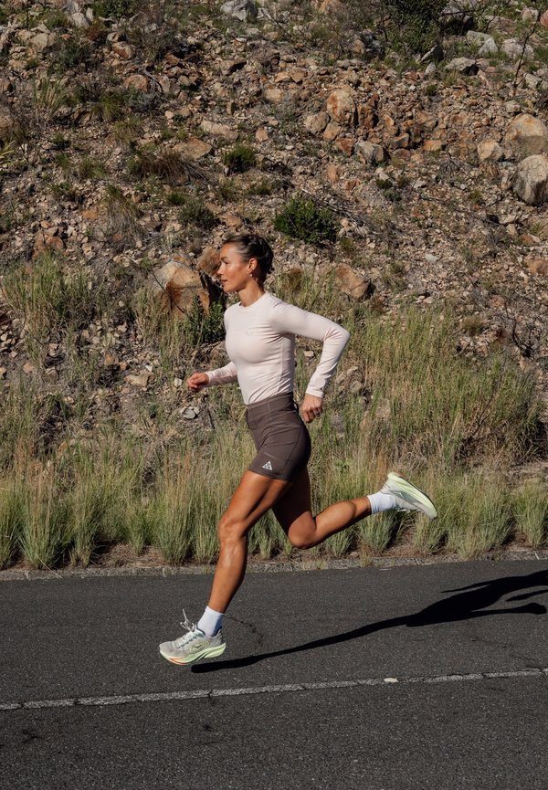 Mujer atlética corriendo por una carretera pavimentada junto a un terreno rocoso con hierba seca, vistiendo una camiseta ligera de manga larga, pantalones cortos oscuros y zapatillas de correr.