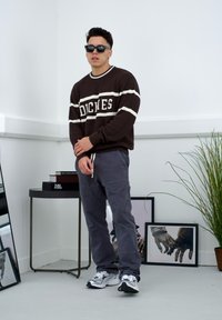 Smeđi rebrasti pulover s bijelim prugama i "DICKIES" logotipom, uparen s običnim sivim hlačama i bijelim tenisicama; sunčane naočale upotpunjuju izgled.