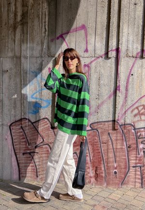 Camisa de rayas negras y verdes de gran tamaño, pantalones crema, zuecos marrones y un bolso negro, todo ello frente a una pared de hormigón cubierta de graffiti.
