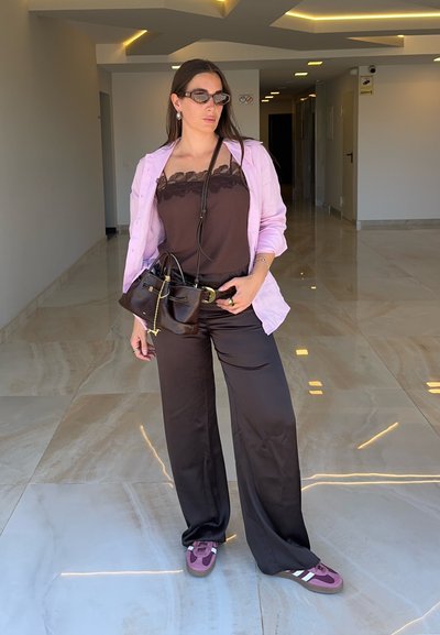 Mujer con gafas de sol, top marrón de encaje, camisa rosa, pantalones negros, zapatillas rosas y sosteniendo un bolso marrón en un pasillo moderno.