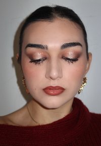 Nærbilde av et ansikt med nøytral sminke; hasselbrune øyne med glitrende øyenskygge, blanke brune lepper og gulløreringer. Strikkegenser i teksturert burgunder.