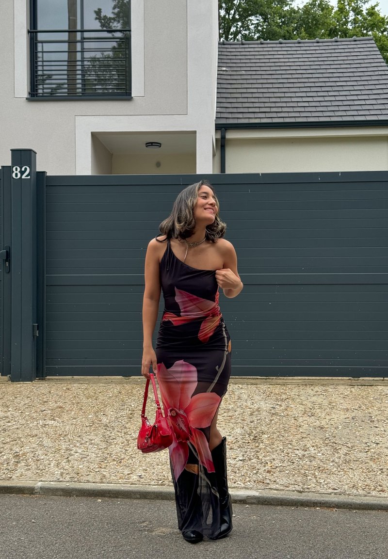 Μαύρο φόρεμα με λουλούδια σε ροζ και πορτοκαλί μοτίβα, εφαρμοστή σιλουέτα, πλαϊνό σκίσιμο, συνδυασμένο με μαύρες μπότες μέχρι το γόνατο και μια μικρή κόκκινη τσάντα.