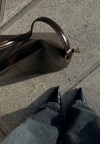 Scarpe nere con punta a punta indossate con jeans scuri accanto a una borsetta in pelle marrone scuro su un pavimento grigio piastrellato.