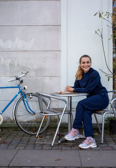 Bicicleta azul aparcada junto a una mesa redonda de metal; una persona con un chándal azul marino se sienta en una silla de metal, con zapatillas de colores.