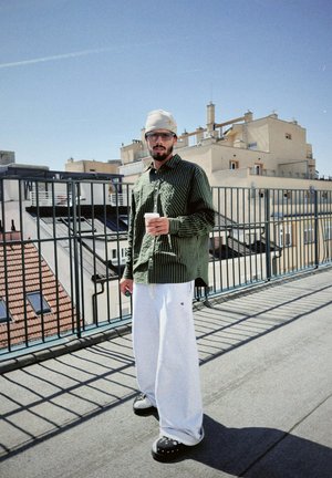 Camisa de botones a rayas verdes, pantalones de chándal grises y zuecos negros. Sosteniendo una taza de café con un tejado y edificios de fondo.