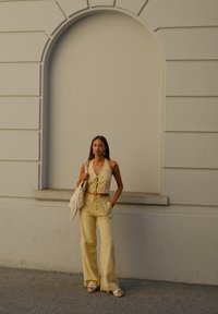 Leinen-Zweiteiler in blassem Gelb, bestehend aus einem Haltertop mit Bindeband vorne und weiten Hosen. Als Accessoires sind eine Fransenhandtasche und Sandalen enthalten.