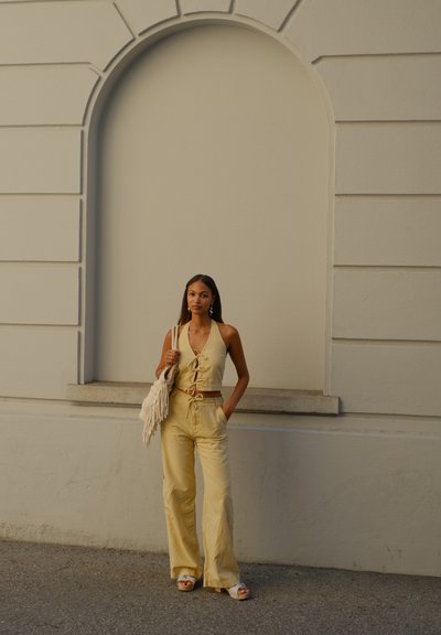 Conjunto de dos piezas de lino en amarillo pálido, que incluye un top halter con lazo en la parte delantera y pantalones de pierna ancha. Los accesorios incluyen un bolso con flecos y sandalias.