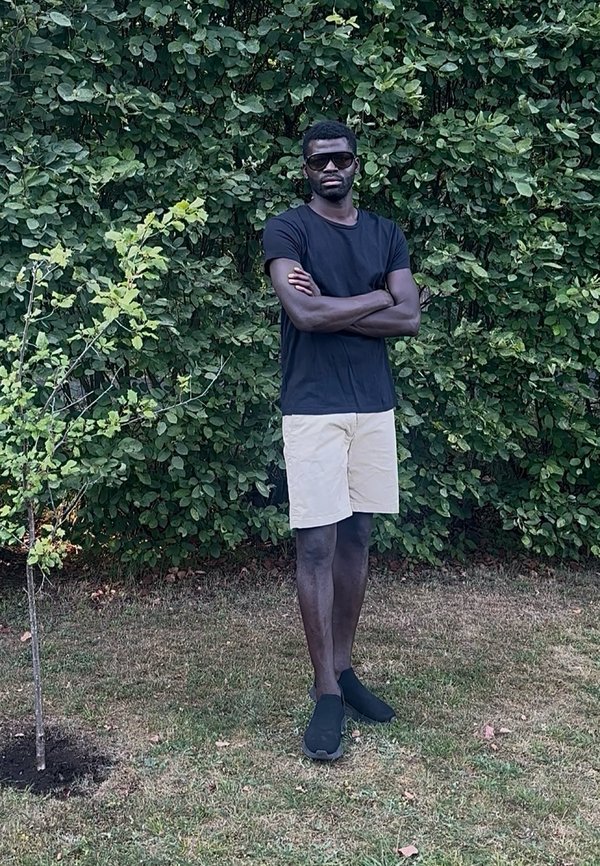 Homme portant des lunettes de soleil, un t-shirt noir, un short beige et des chaussures noires, debout, les bras croisés, sur l'herbe près de buissons verts feuillus.