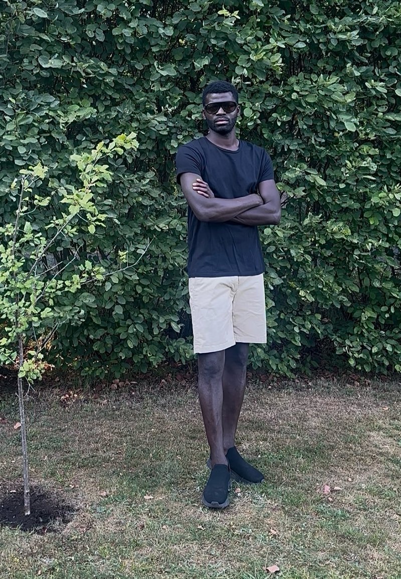 Homme portant des lunettes de soleil, un t-shirt noir, un short beige et des chaussures noires, debout les bras croisés sur l'herbe près de buissons verts feuillus.