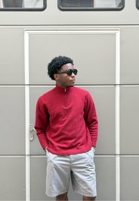 Pulôver de malha vermelho com gola alta, fecho de zíper à frente; calções cinza claro; óculos de sol pretos; de pé contra um fundo cinza texturizado.