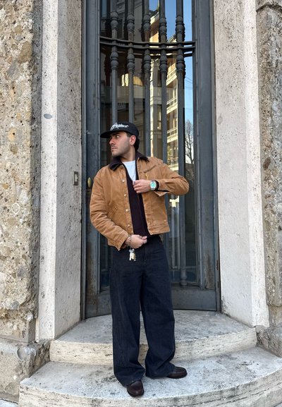 Chaqueta de pana marrón con cuello negro, camiseta negra, vaqueros negros holgados, zapatos marrones y gorra negra. Llaveros visibles en la mano.