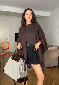 Mlada žena s kovrčavom kosom nosi smešani smešani šal i crne šortse, držeći veliku bež i smeđu tašnu u dnevnoj sobi.
