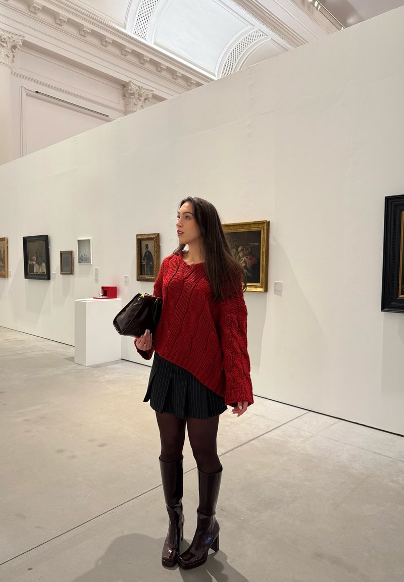 Femme en pull rouge, jupe noire et bottes, tenant un sac à main, debout dans une galerie avec des peintures encadrées sur des murs blancs.