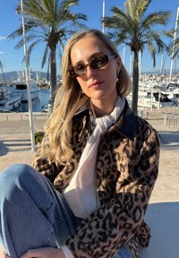 Veste à motif léopard avec un col noir, un pull gris clair et un jean bleu. Le décor comprend des palmiers et des yachts en arrière-plan.