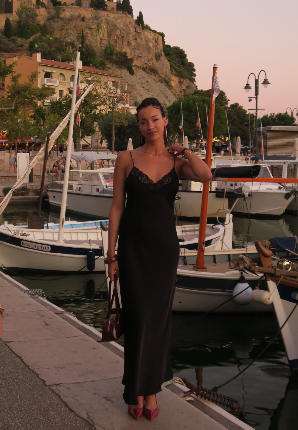 Vestido maxi de cetim preto com acabamento de renda, alças de espaguete e silhueta ajustada. Acessórios incluem uma bolsa burgundy e sapatos, com fundo à beira-mar.