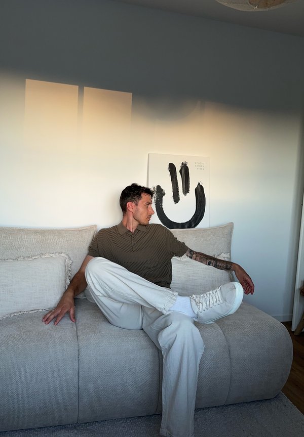 Man met een getatoeëerde arm, gekleed in een bruin shirt en witte broek, zit op een lichtgrijze bank in een minimalistisch ingerichte kamer met kunst van een lachend gezicht.