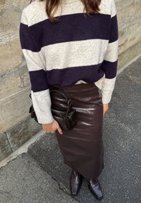 Donna che indossa un maglione a strisce bianco e nero, una gonna di pelle marrone scuro, stivali abbinati e tiene in mano una piccola borsa a tracolla di pelle nera.