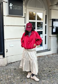 Rød jakke med lynlås og hvide detaljer, grå rullekrave, beige lagdelt nederdel og beige sneakers. Baggrunden viser en bygningsindgang.