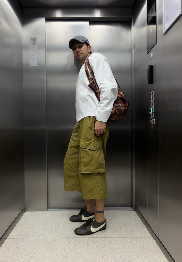 Hombre que lleva una camisa blanca de manga larga, pantalones cortos cargo verdes, zapatillas negras y gorra, cargando una bolsa a cuadros, de pie dentro de un ascensor de acero inoxidable.