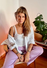 Weißes Button-Down-Hemd über einem enganliegenden weißen Tanktop, kombiniert mit helllila gestreiften Jogginghosen. Lange Haare, sichtbare Tattoos an den Armen.