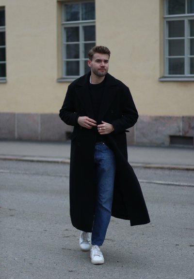 Abrigo largo negro, suéter negro de punto, jeans azules, zapatillas blancas. Entorno al aire libre con un edificio de color claro y ventanas al fondo.