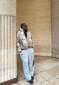 Pufasta jakna u bež boji s zelenim uzorcima, uparena s sivim trenirkama s crnom zvijezdom. Stojeći ispred teksturiranog kamenog zida.