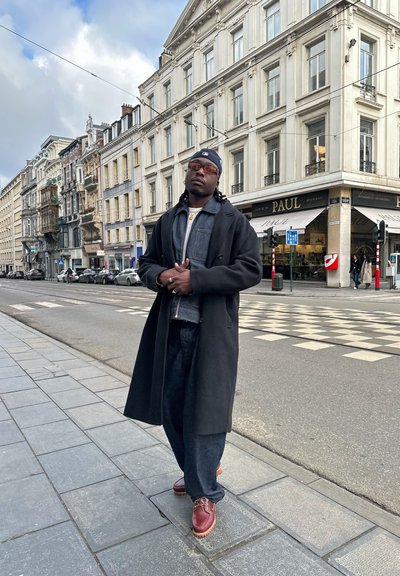 Abrigo negro, chaqueta de mezclilla gris, pantalones oscuros de pierna ancha, zapatos marrones con cordones. Escena urbana con una arquitectura clásica de fondo.