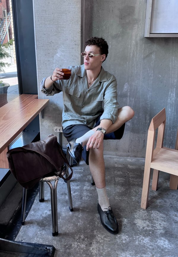 Jeune homme portant des lunettes de soleil, une chemise vert clair et un short noir, assis en tailleur, sirote un café dans un café moderne aux murs en béton et au mobilier en bois.