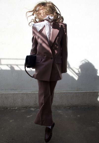 Mujer con traje pantalón malva y blusa blanca salta con el cabello cubriendo su rostro, sosteniendo un bolso negro frente a una pared blanca con sombras.